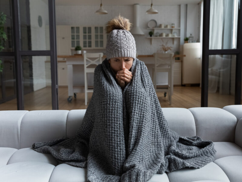 person wrapped in a gray blanket in a home that is still cold even with the heat on caused by drafts, poor insulation, and HVAC issues.