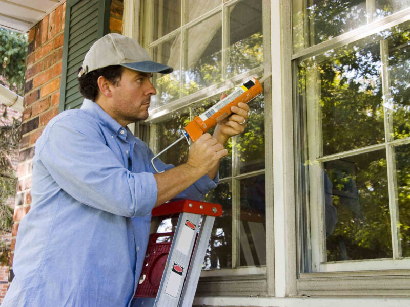 dark-haired man in a blue button-down shirt and ball cap on a ladder caulking the exterior of a window