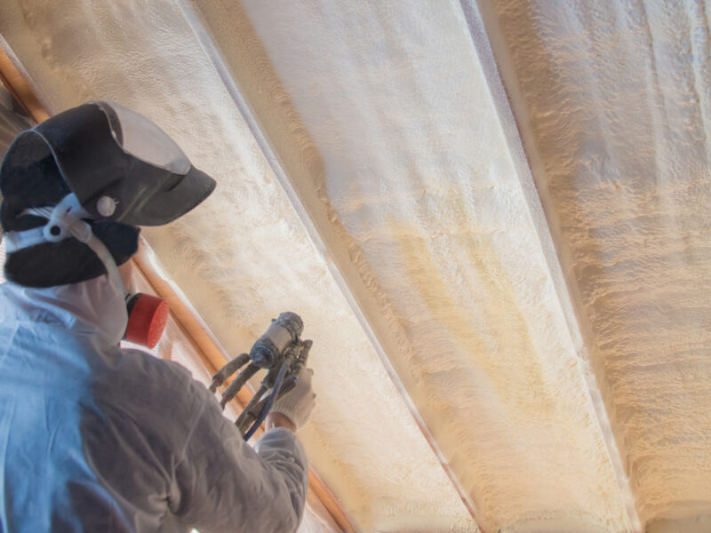 person in white protective gear and a black helmet installing spray foam insulation on a ceiling