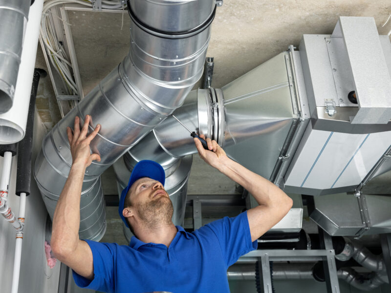 man in a blue short-sleeve shirt and hat working on overhead air duct pipe
