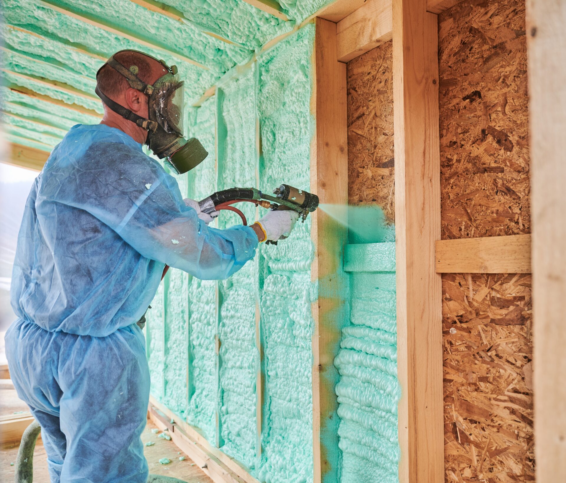 technician in a blue protective suit applying spray foam insulation to a wall