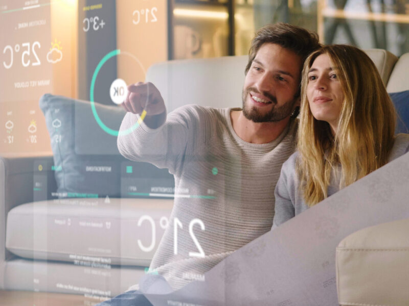 dark-haired bearded man and long-haired woman sit on a couch using smart home controls