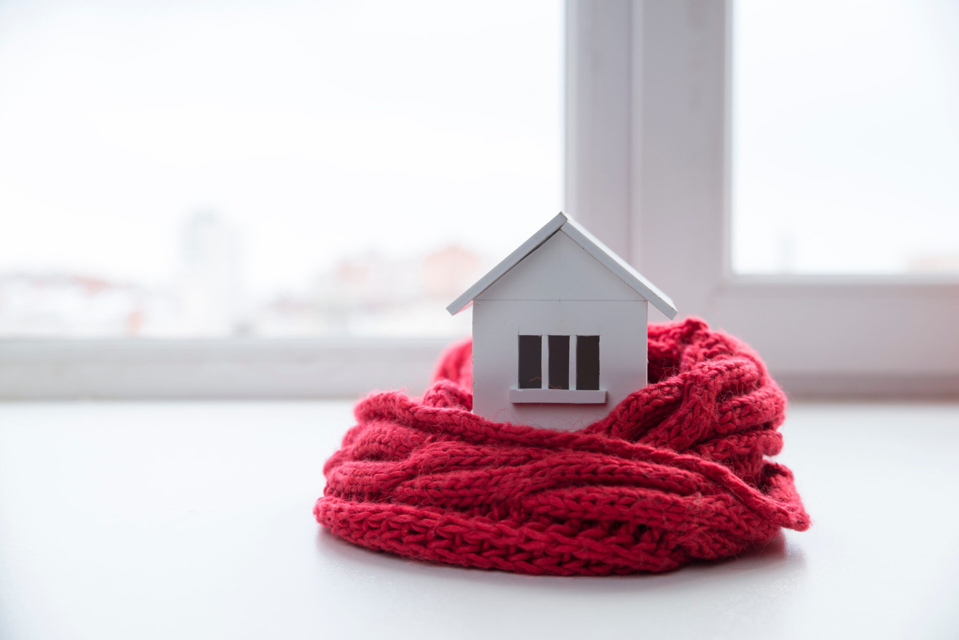 model of a white house with three windows wrapped in a red knit scarf on a white tabletop with a window in the background