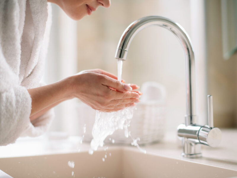 woman in a white robe cupping hot water in her hands from a silver faucet after installing a Bradford white hot water heater