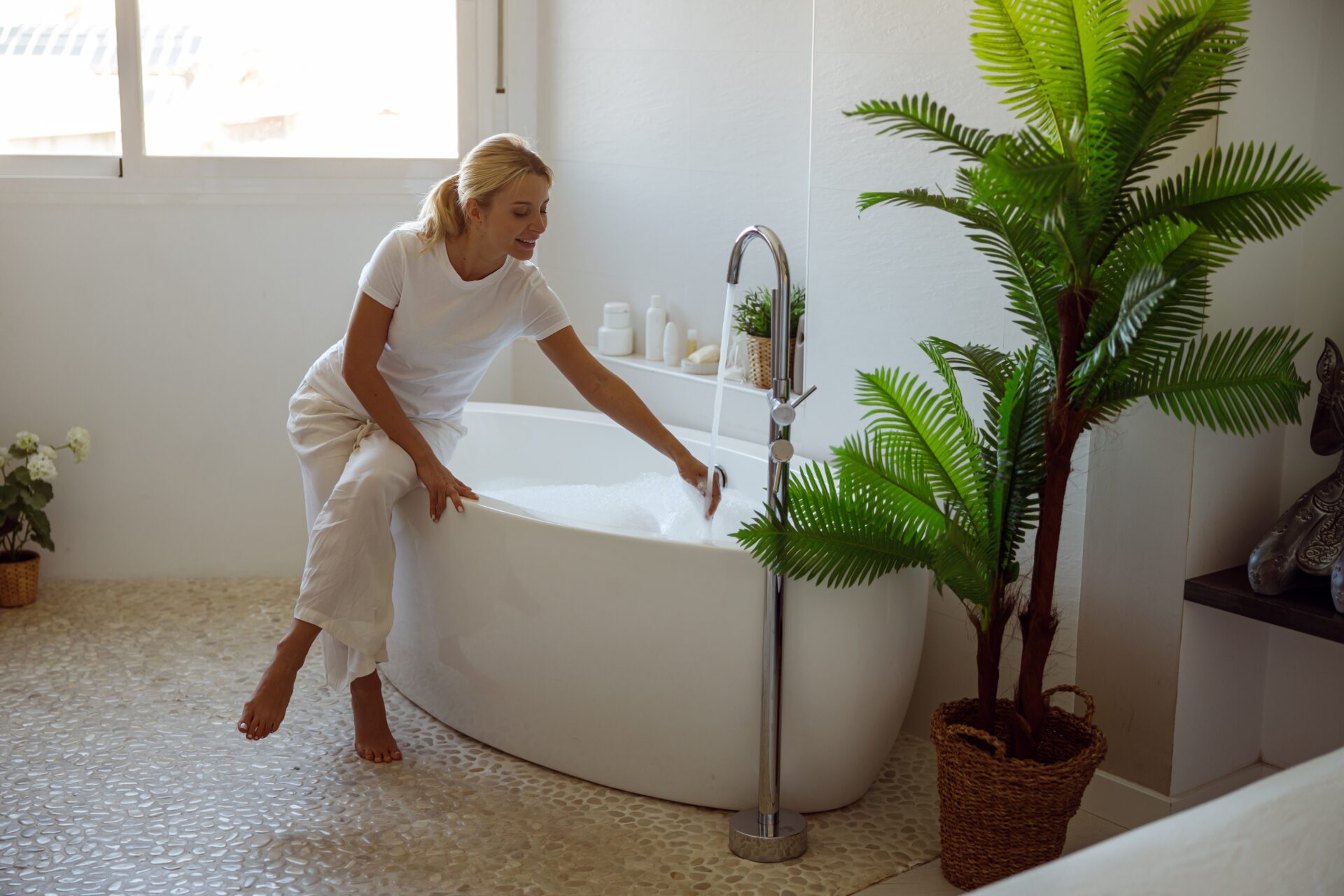 A blond woman wearing a white shirt and pants draws a bath, happy with the temperature of the water