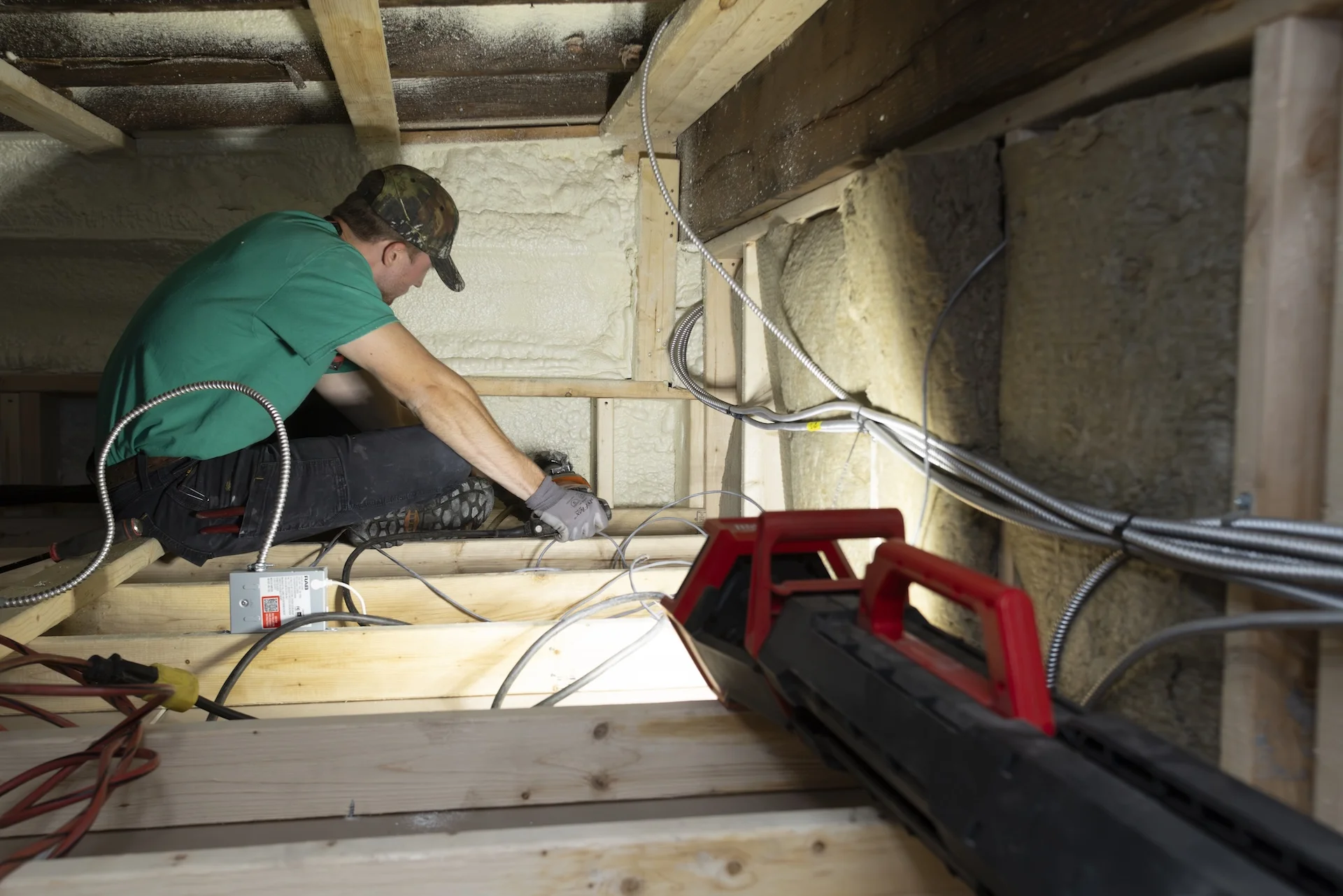 Technician in a green shirt and work gloves crouched in an insulated attic or crawlspace, working on wiring between exposed wooden ceiling joists with a red work light in the foreground.